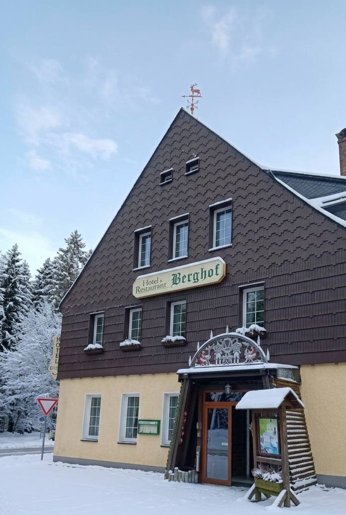 Hotel‑Restaurant ›Berghof‹ mit brauner Schindelfassade und verschneitem Vorplatz